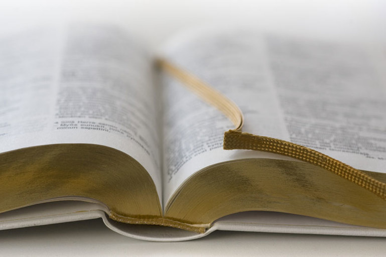 Valkoinen vihkiraamattu lepää avoimena valkoisella pöydällä. Raamatussa on kultainen kirjamerkki.