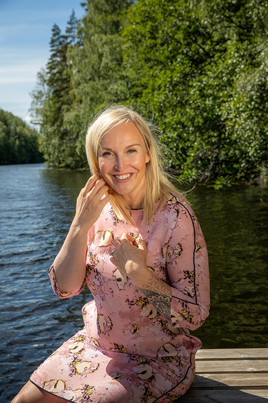 Jutta Larm istuu laiturilla. Taustalla järvi ja vehreää metsää. Aurinkoinen sää.