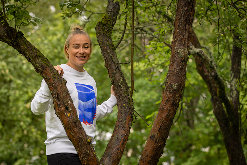 Ella Räsänen kurkistaa hymyillen puunrunkojen takaa. Taustalla vehreää metsää.