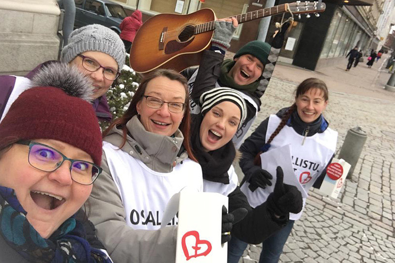 Kuusi henkilö yhteisvastuuliivit päällään ja lippaat kädessään kokoontuneet yhteen iloisen oloisesti.