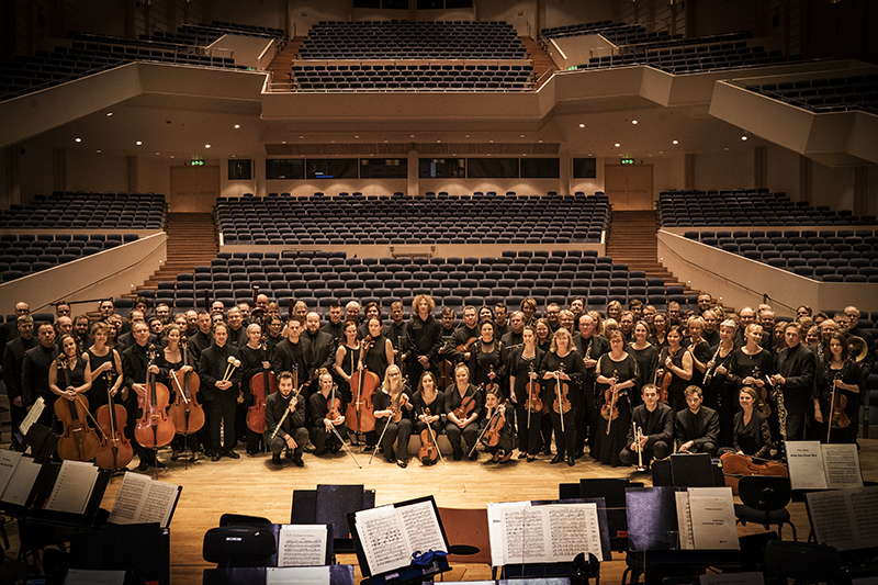 Tampere Filharmonia soi nyt sosiaalisessa mediassa. Filharmonian jäsenet poseeravat suuressa ryhmäkuvassa kameralle. Sali on muuten tyhjä ja sävyt ovat lämpimät.