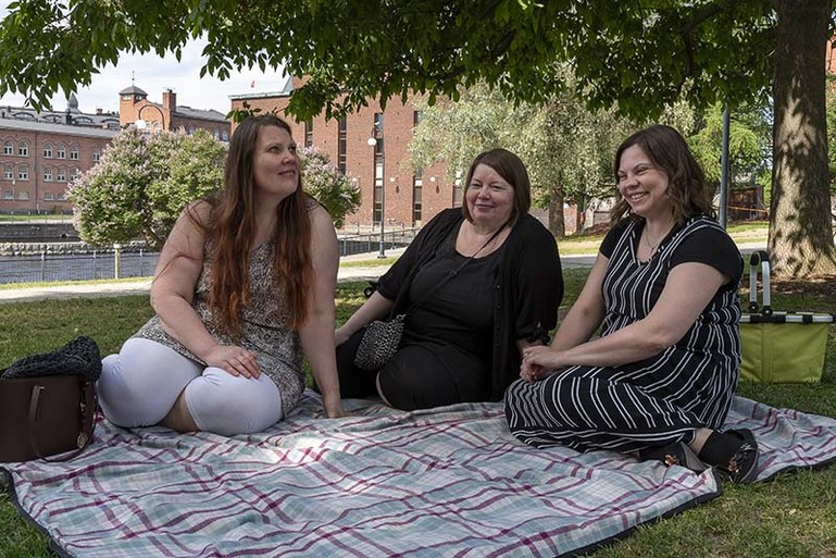 Kolme henkilöä istuu viltillä nurmikolla. Taustalla on koskimaisema.