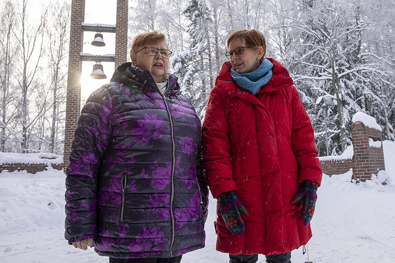 Kaksi naista seisovat talvisessa säässä Hervannan kirkon ovilla. Takana näky kellotorni, joiden takaa aurinko paistaa.