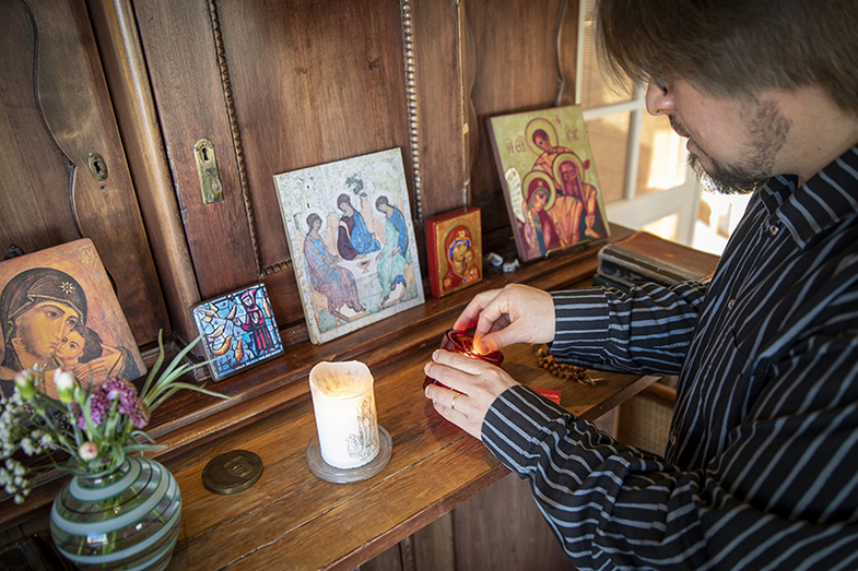 Mies raidallisessa kauluspaidassa sytyttää kotialttarille kynttilää. Alttarilla on myös näytillä viisi ikonia.