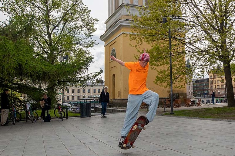 Oranssipaitainen poika skeittaa Vanhan kirjastotalon edessä. Kaverit seuraavat sivussa.