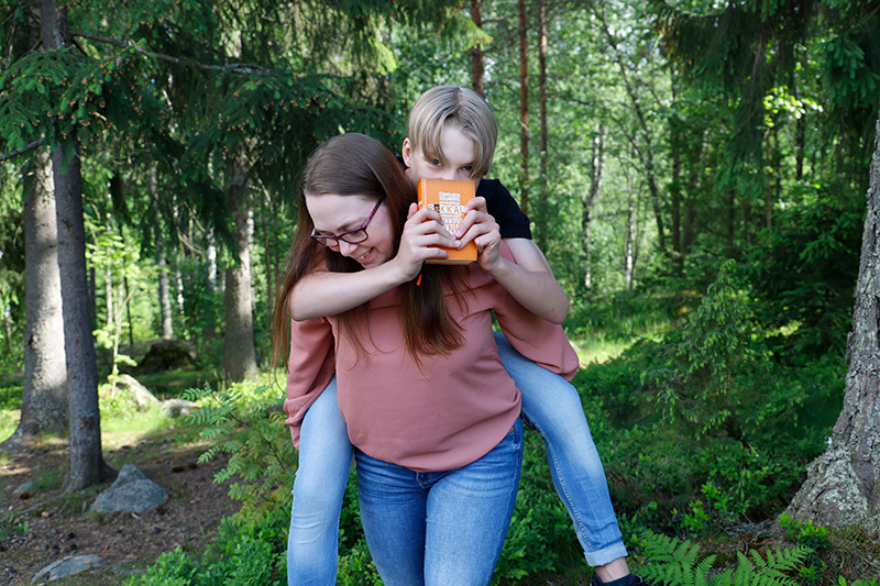 Äiti pitää reppuselässä rippikouluikäistä poikaansa metsässä. Pojan kädessä oranssikantinen raamattu.