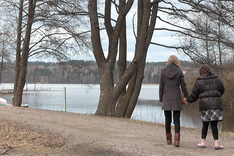 Syksyisellä hiekkatiellä järven rannan tuntumassa kävelee kaksi naista selin kameraan päin pitäen toisiaan kädestä kiinni.