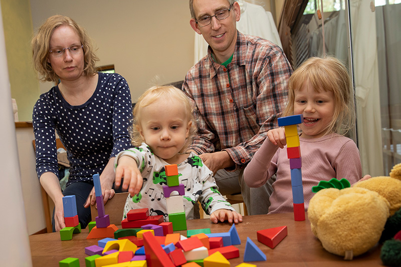 Edustalla kaksi lasta leikkii värikkäillä puupalikoilla pöydän ääressä. Vanhemmat istuvat taustalla ja katsovat lasten leikkejä.