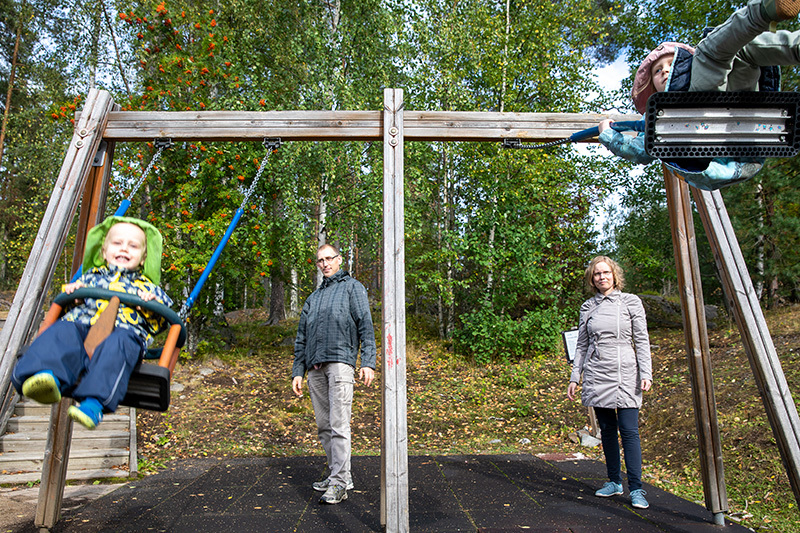 Kaksi lasta keinuu keinuissa. Vanhemmat seisovat takana ja antavat keinuille vauhtia.