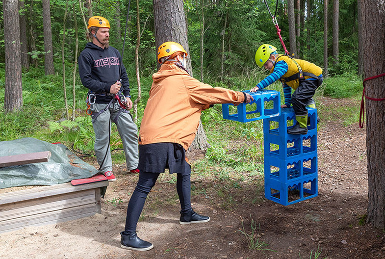 Poika kiipeilee valjaiden varassa köysiradalla ulkona, jalkojen alla torni sinisiä muovilaatikoita. Aikuinen auttamassa kädestä pitäen ja taustalla toinen aikuinen.