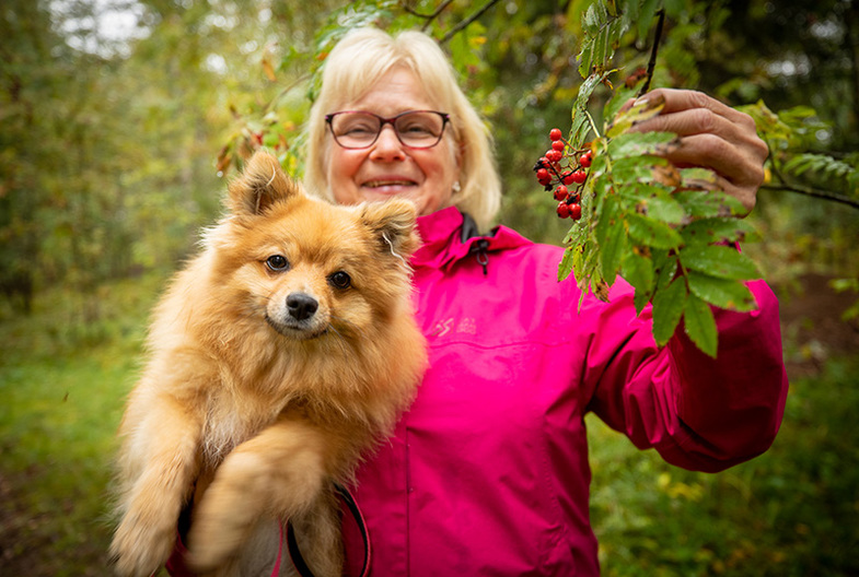 Nainen hymyilee koira sylissään ja pitää toisella kädellä kiinni pihlajan oksasta.