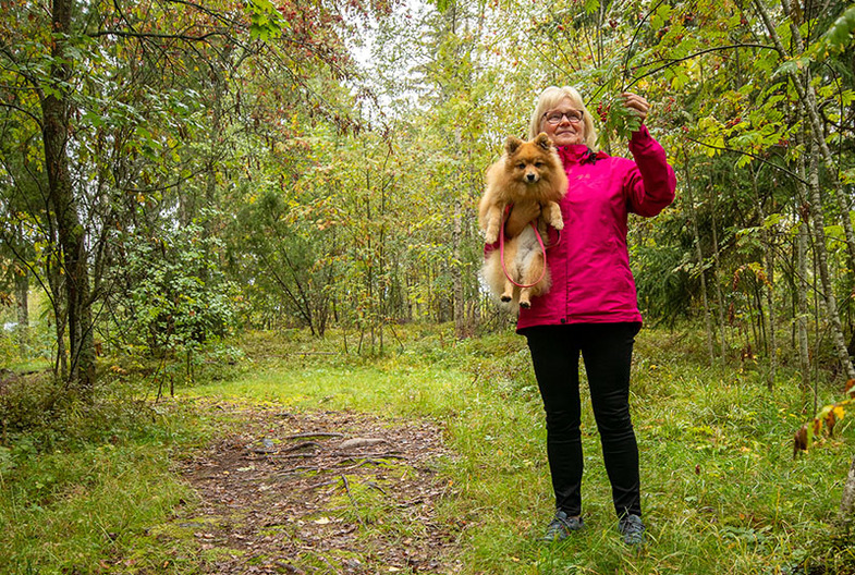 Nainen metsäpolulla, koira sylissään. Pitää toisellä kädellä kiinni pihlajan oksasta.