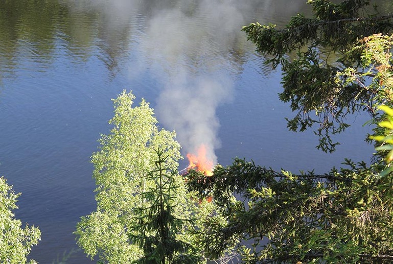 Järven rannassa näkyy puiden takaa liekkejä ja savua.