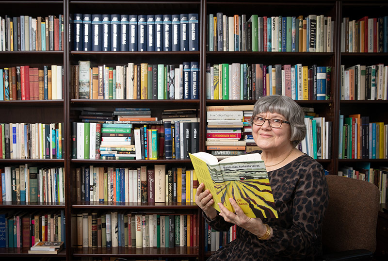 Nainen käsissään kirja, taustalla kirjahylly, jossa on paljon kirjoja.