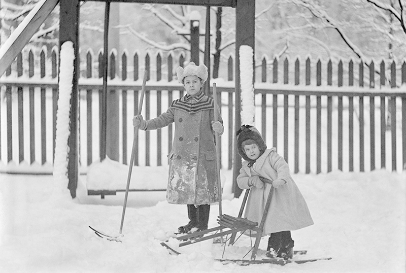 1900-luvun alkupuolen mustavalkoisessa kuvassa kaksi tyttöä talvisissa puuhissa. Toinen hiihtää ja toinen potkukelkkailee.