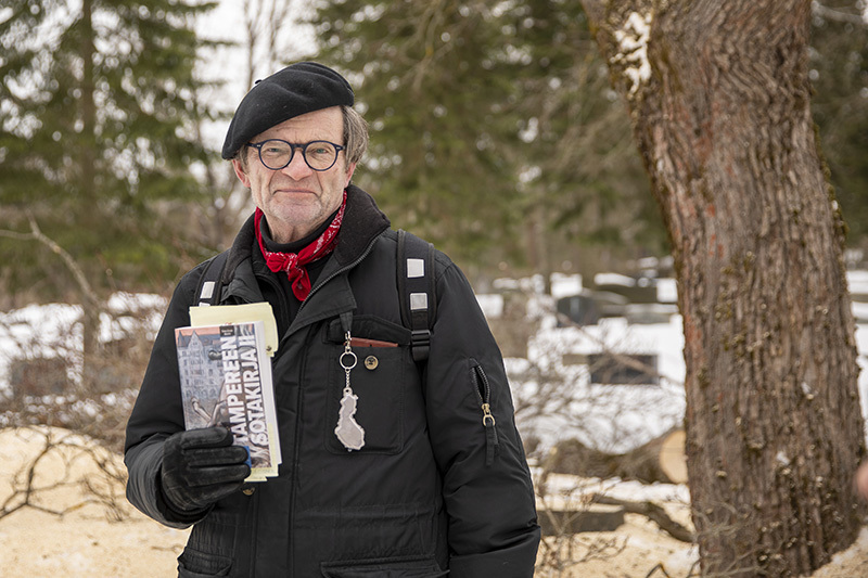 Talvinen kuva miehestä hautausmaalla kirja kädessään. Vieressä on puunrunko.