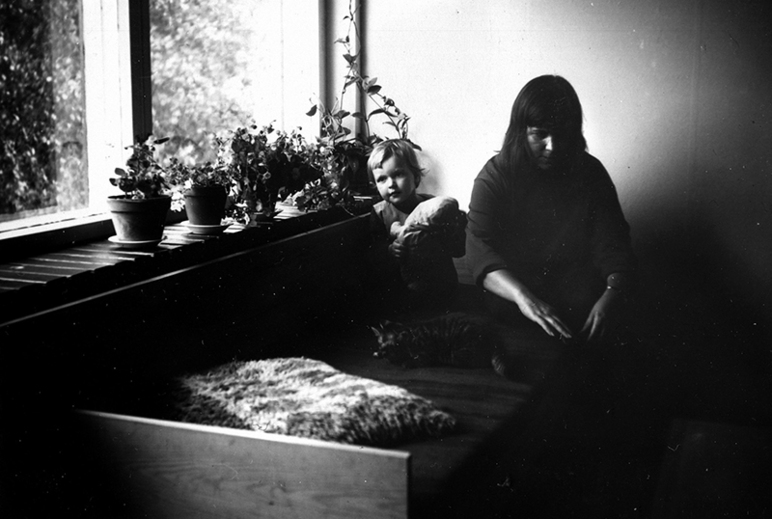 Tummahko kuva sängyllä istuvasta naisesta ja ikkunan luona olevasta pikkupojasta. Ikkunalaudalla on huonekasveja ruukuissa.