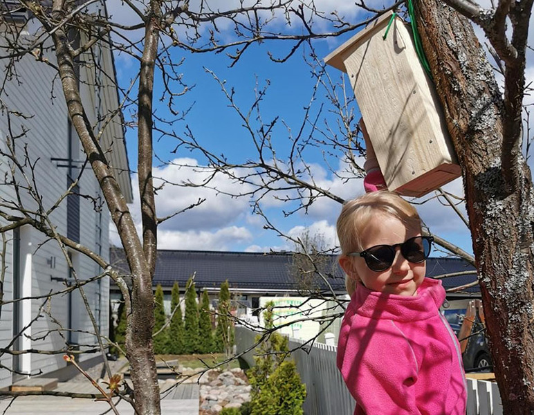 Tyttö aurinkolasit silmillään näyttää puussa olevaa linnunpönttöä. Taustalla on rakennuksen nurkka ja pihamaata.