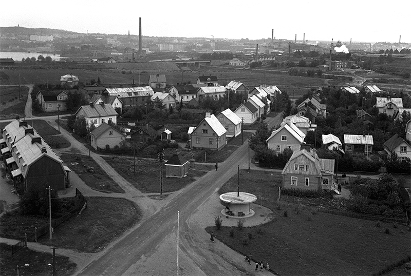 Mustavalkoinen kuva 1930-luvulta. Puutaloja ja hiekkateitä. Taustalla tehtaiden piippuja ja kaupunki.