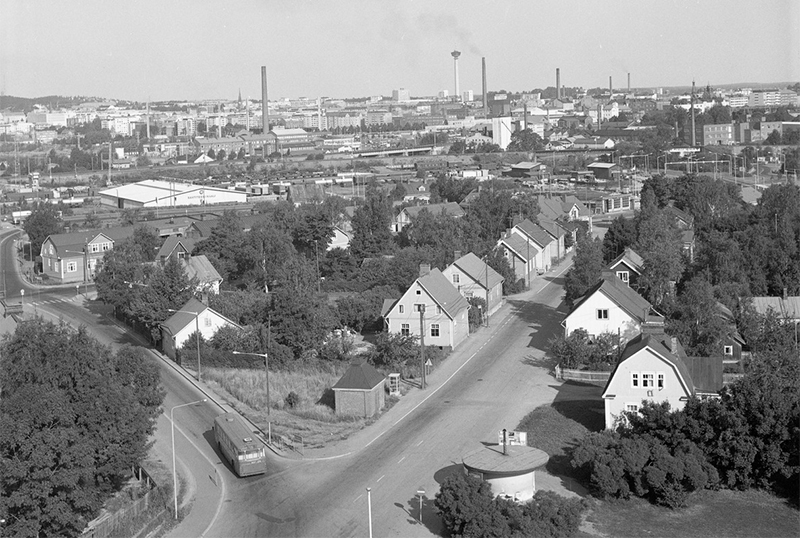 Mustavalkoinen kuva, jossa edustalla puisia omakotitaloja. Taustalla kaupunki tehtaan piippuineen.