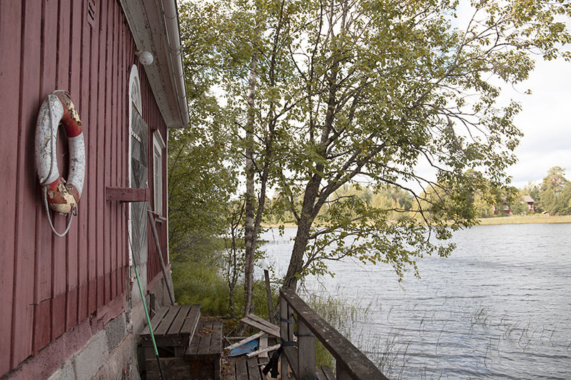 Vasemmalla näkyy punaista rakennusta, jonka seinällä on vanha pelastusrengas. Oikealla on järveä, puu ja laituri.