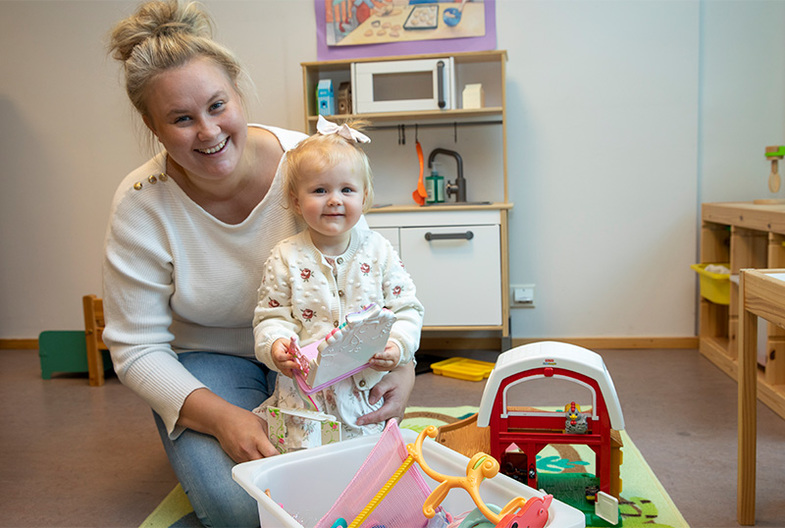 Nainen hymyilee lapsen kanssa kameralle lelukorin takaa.