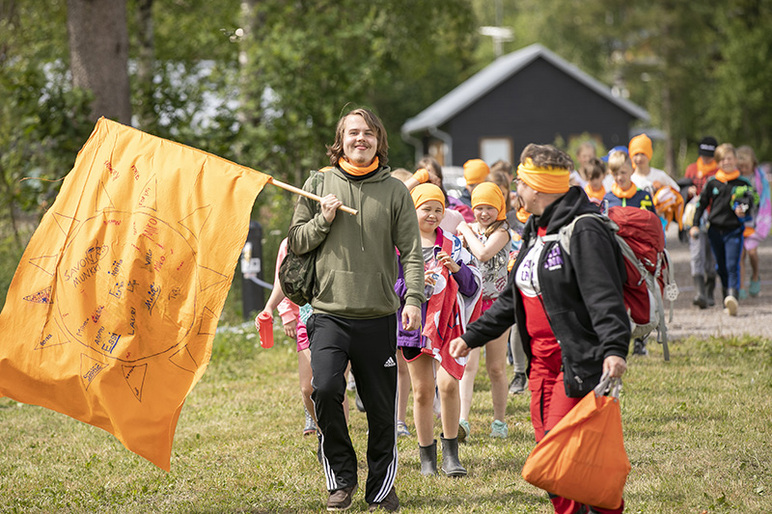 Nuori ihminen kantaa lippua edellä ja iloiset lapset seuraavat perässä.