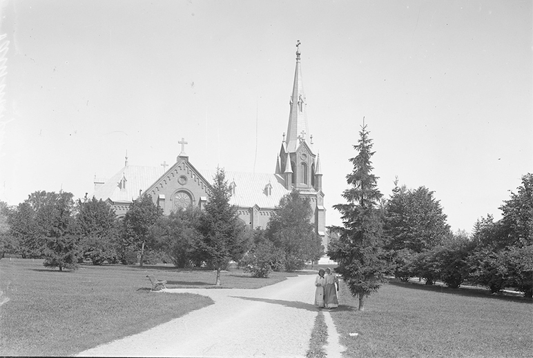 Mustavalkoinen kuva vuodelta 1908. Taustalla kirkko ja puistossa puun juurella kaksi ihmistä suojassa auringolta.