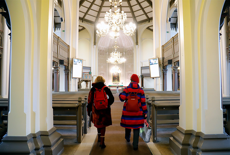 Kaksi naista kävelee kirkon käytävää pitkin ulkovaatetuksessa.