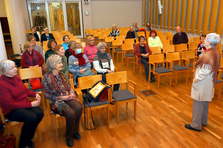 Nainen puhuu mikrofooniin ja hänen edessään on yleisöä, jotka istuvat tuoleilla.