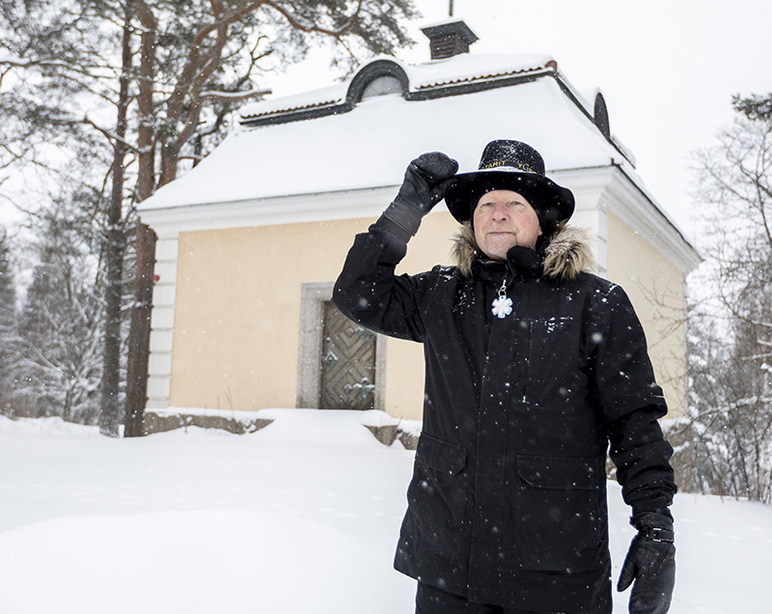 Etualalla mies koskettaa hattua, jossa on tekstiä. Taustalla on puinen neliömäinen rakennus, jonka ovessa ja katossa on monia yksityiskohtia.