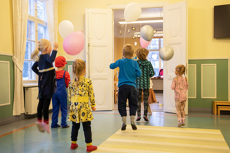 Lapset pomppivat selin kameraan oviaukon edessä ja yrittävät kiskoa ilmapallojen naruista.