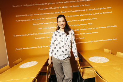 Hymyilevä nainen seisoo pöytien välissä ja pitää kiinni tuolin selkänojasta. Taustalla on tekstiä taulussa.