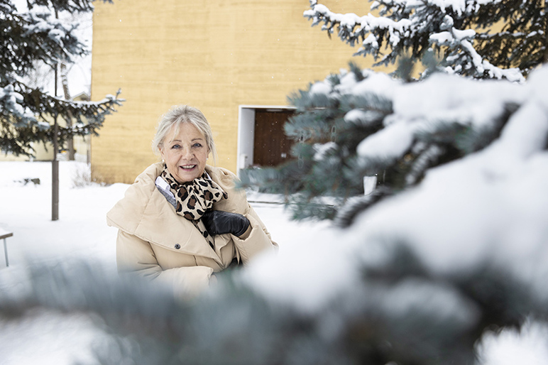 Hymyilevä nainen lumisessa maisemassa kuusenoksan vieressä. Taustalla näkyy vaaleaa seinää ja ovi.