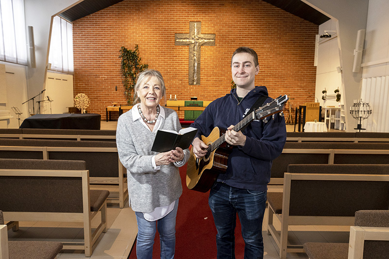 Nainen kirja ja mies kitara kädessään seisovat hymyillen kirkon käytävällä. Takana näkyy alttari ja seinällä iso risti.