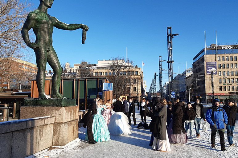 Talvisella kadulla on väkeä pitkähelmaisissa mekoissa ja tummissa juhlapuvuissa. Jotkut ottavat valokuvia. Vasemmalla näkyy patsas.