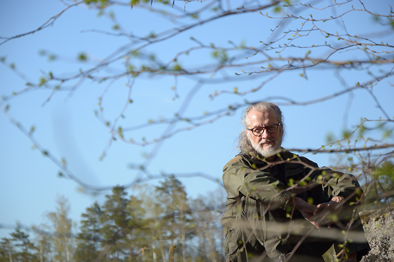 Silmälasipäinen mies, jolla on parta, istuu luonnossa. Puissa on silmuja.