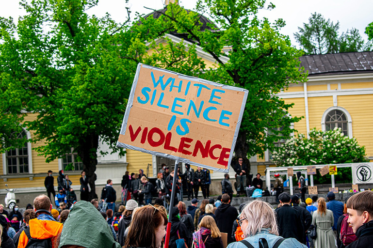 Suuri väkijoukko ulkona Tampereen keskustorilla. Väkijoukossa yhdellä on suuri kyltti, jossa lukee englanniksi: White silence is violence.