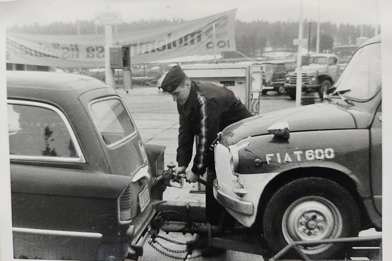 Vanha kuva autoa tankkaavasta miehestä kahden auton keskellä.