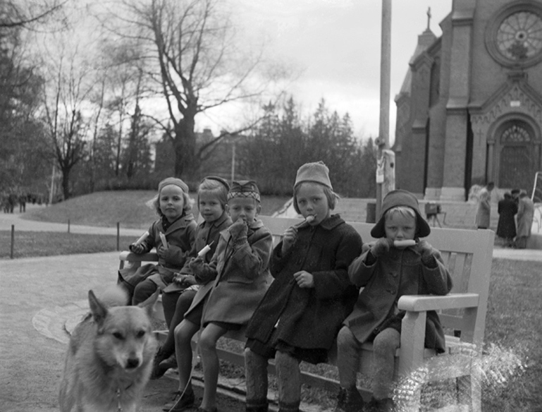 Vanha kuva viidestä lapsesta, jotka istuvat penkillä syömässä jäätelöpuikkoja. Edessä on koira ja taustalla kirkko.