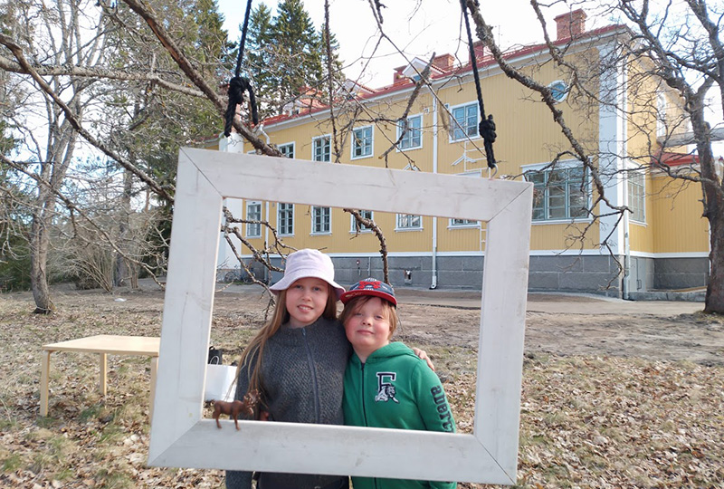 Kaksi lasta pihalla katselee ison tyhjän kehyksen läpi. Taustalla on iso puurakennus.