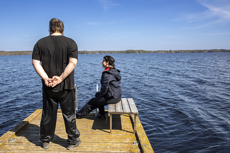 Laiturilla on kaksi ihmistä, joista toinen seisoo selin ja toinen istuu sivuttain katsojaan.