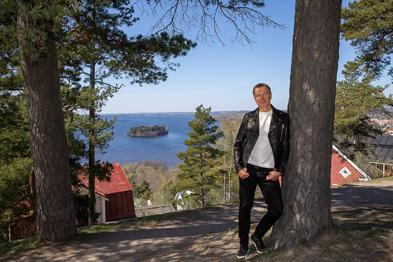 Hymyilevä mies nojaa rennosti puuhun maisemassa, jossa taustalla on taloja ja vesistöä.