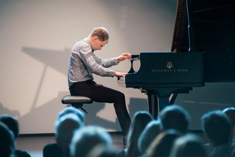 Sivuprofiili pianoa soittavasta henkilöstä. Kuvan etualalla on selin kameraan olevaa yleisöä.