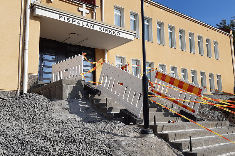 Portaikon vierellä on soraa ja sen reunoilla remonttiaitoja ja nauhoja. Portaikko vie pitkänomaiseen rakennukseen. Portaikon yläpäässä olevassa sadelipassa on teksti Pispalan kirkko ja sen päällä on risti.