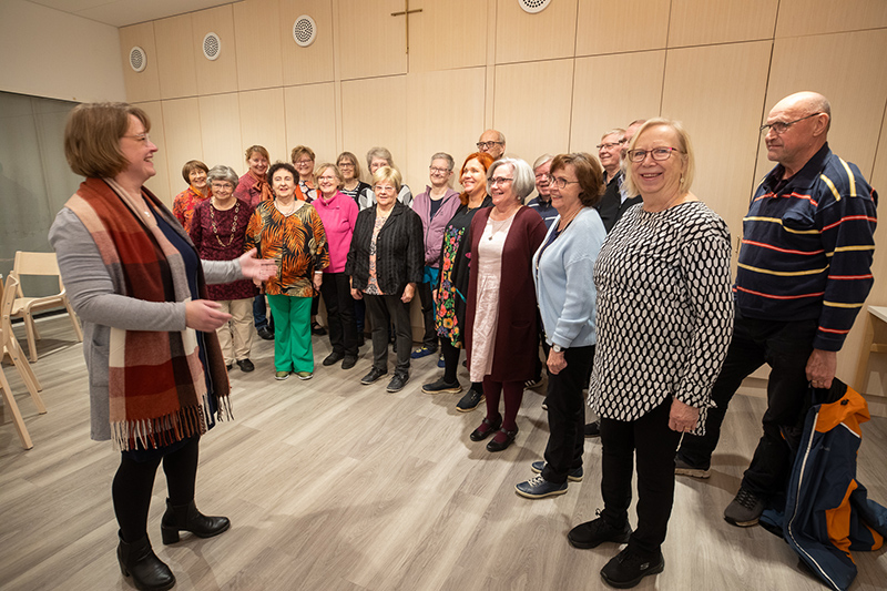 Joukko iloisia ihmisiä seisoo puisessa salissa, jonka seinällä on risti. Vasemmalla puolella erillään muista seisova nainen heiluttaa käsiään.