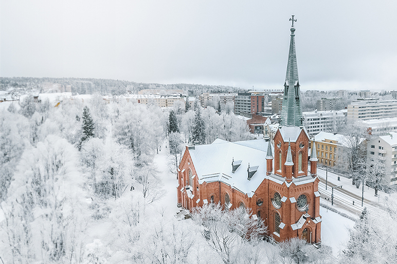 Kirkko, jossa on korkea torni, lumisessa maisemassa kuvattuna ylhäältä sivusuunnasta. Edessä on puita ja taustalla kerrostaloja.