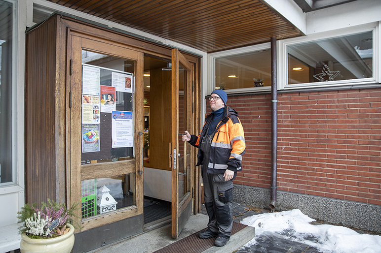 Työvaatteisiin puettu henkilö pitää rakennuksen puukehyksistä ovea raollaan.