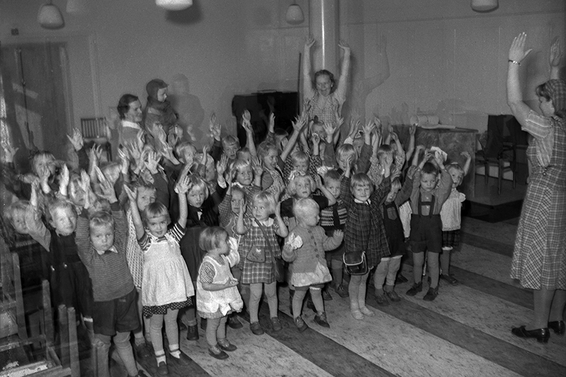 Vanhassa valokuvassa lapset seisovat kädet pystyssä seurakuntien kerhossa.