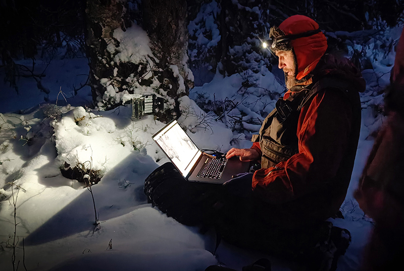 Polvillaan oleva henkilö tutkii tietokonetta otsalampun valossa pimeässä talvimetsässä.